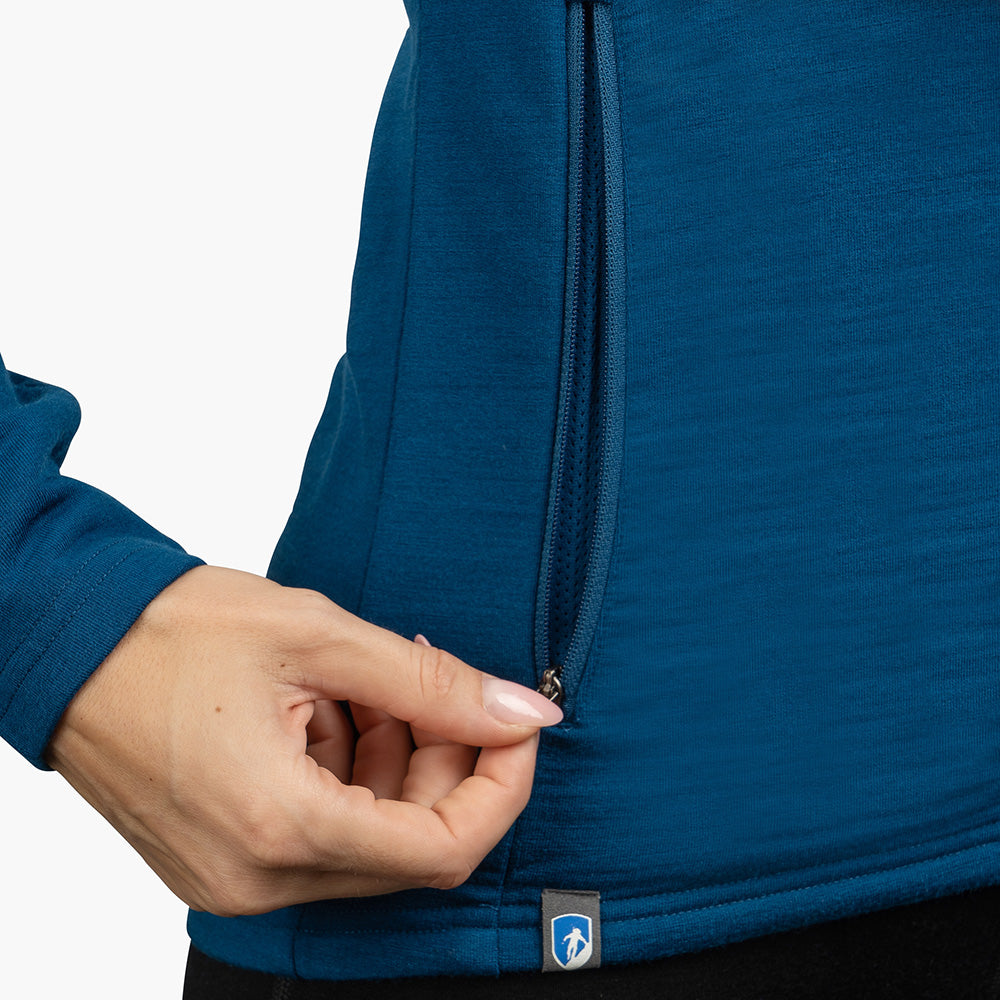 Nahaufnahme einer Hand, die den Reißverschluss einer Merino Jacke Damen 270 g/m2 mit Daumenloch und Logoetikett am Saum öffnet.