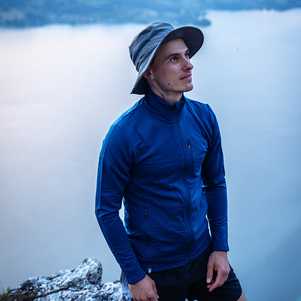 Mann in blauer Merino Jacke Herren 270 g/m² steht auf Felsvorsprung, blickt aufs Wasser. Perfekt für Outdoor-Abenteuer, bietet Wärme, Atmungsaktivität und Flexibilität.