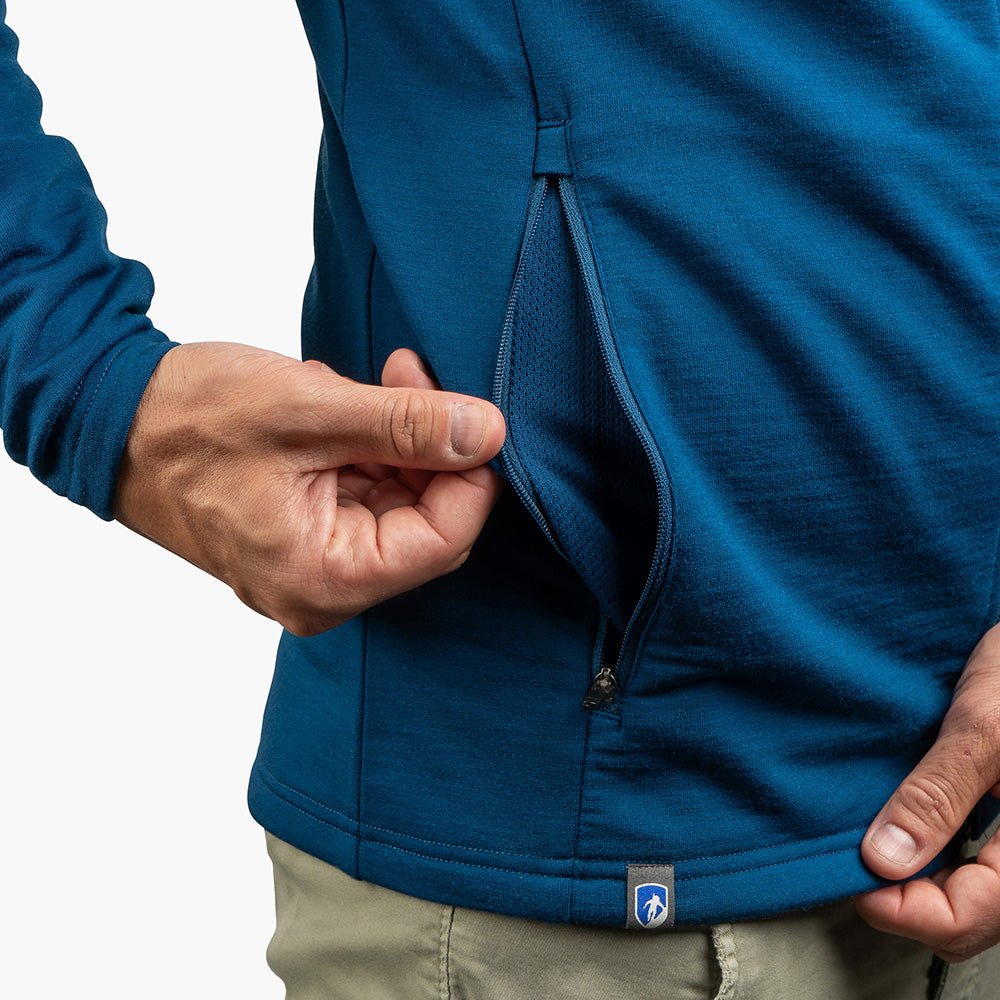Person öffnet seitliche Reißverschlusstasche einer Merino Jacke Herren 270 g/m², zeigt Mesh-Innenfutter. Kleine Logolasche am Saum, kombiniert mit khakifarbenen Hosen.