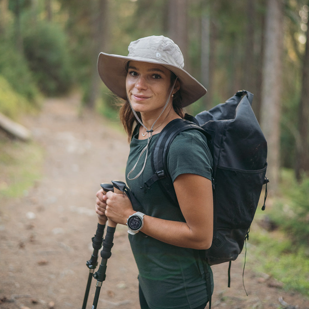 Junge Frau mit Sonnenhut mit UV Schutz Unisex auf Waldweg, bereit zum Wandern, trägt Trekkingstöcke und Smartwatch, verkörpert Abenteuergeist von alpinloacker.com.