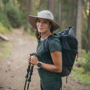 Junge Frau mit Sonnenhut mit UV Schutz Unisex auf Waldweg, bereit zum Wandern, trägt Trekkingstöcke und Smartwatch, verkörpert Abenteuergeist von alpinloacker.com.