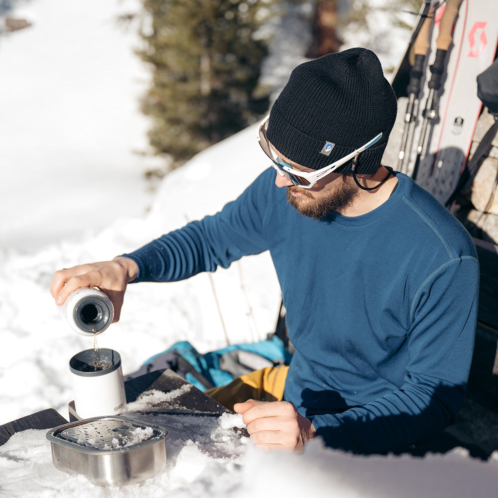 Mann im Schnee mit Merino Langarmshirt Herren 230 g/m², beim Skifahren; er genießt eine Pause, während er ein heißes Getränk aus einem Thermosbecher einschenkt.