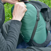 Person verstaut SYN PRO - Synthetik Sommer Schlafsack ultraleicht im Kompressionssack in Seitentasche eines schwarzen Wanderrucksacks; ideal für Outdoor-Abenteuer und nachhaltigen Komfort.