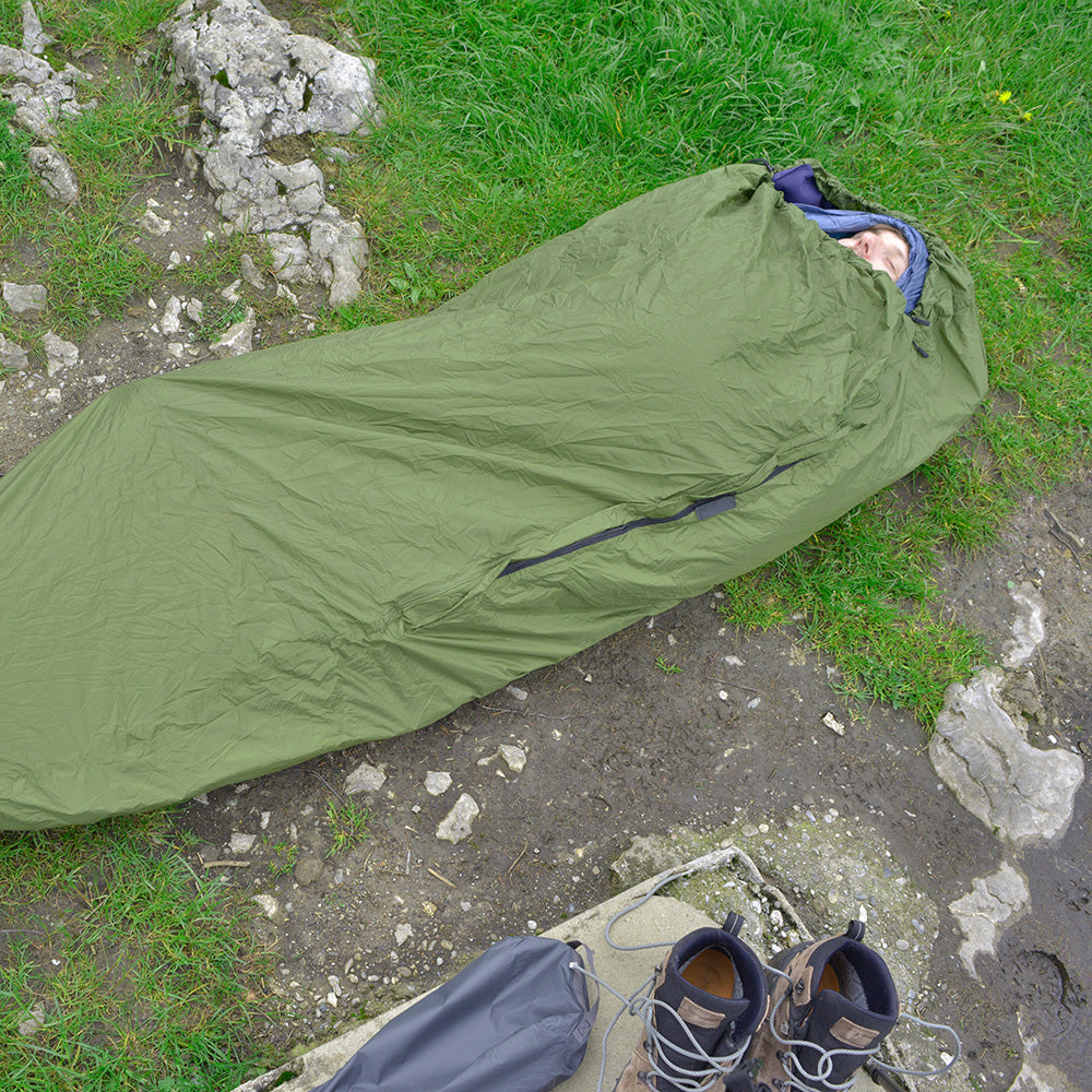 Person im Bivi PRO - leichter Biwaksack auf felsigem Untergrund, mit Wanderstiefeln und Packsack daneben, ideal für Outdoor-Abenteuer und Wetterschutz.