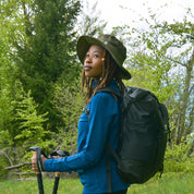 Person mit Sonnenhut mit UV Schutz Unisex, Trekkingstöcken und Rucksack im Wald. Idealer Begleiter für Outdoor-Aktivitäten, leicht und nachhaltig aus recyceltem Material.