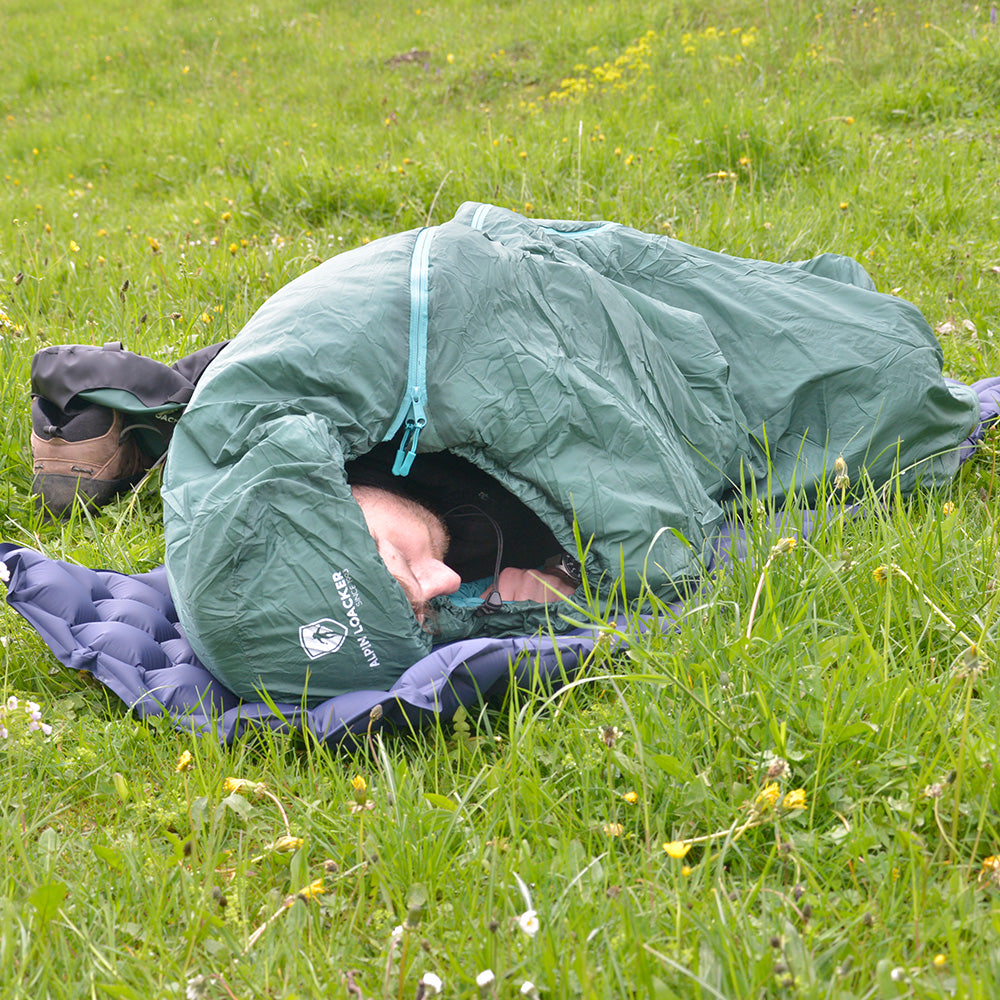 Person in grünem SYN PRO Synthetik Sommer Schlafsack auf Wiese, neben Wanderschuhen. Ideal für Outdoor-Aktivitäten, bietet Komfort und Nachhaltigkeit.