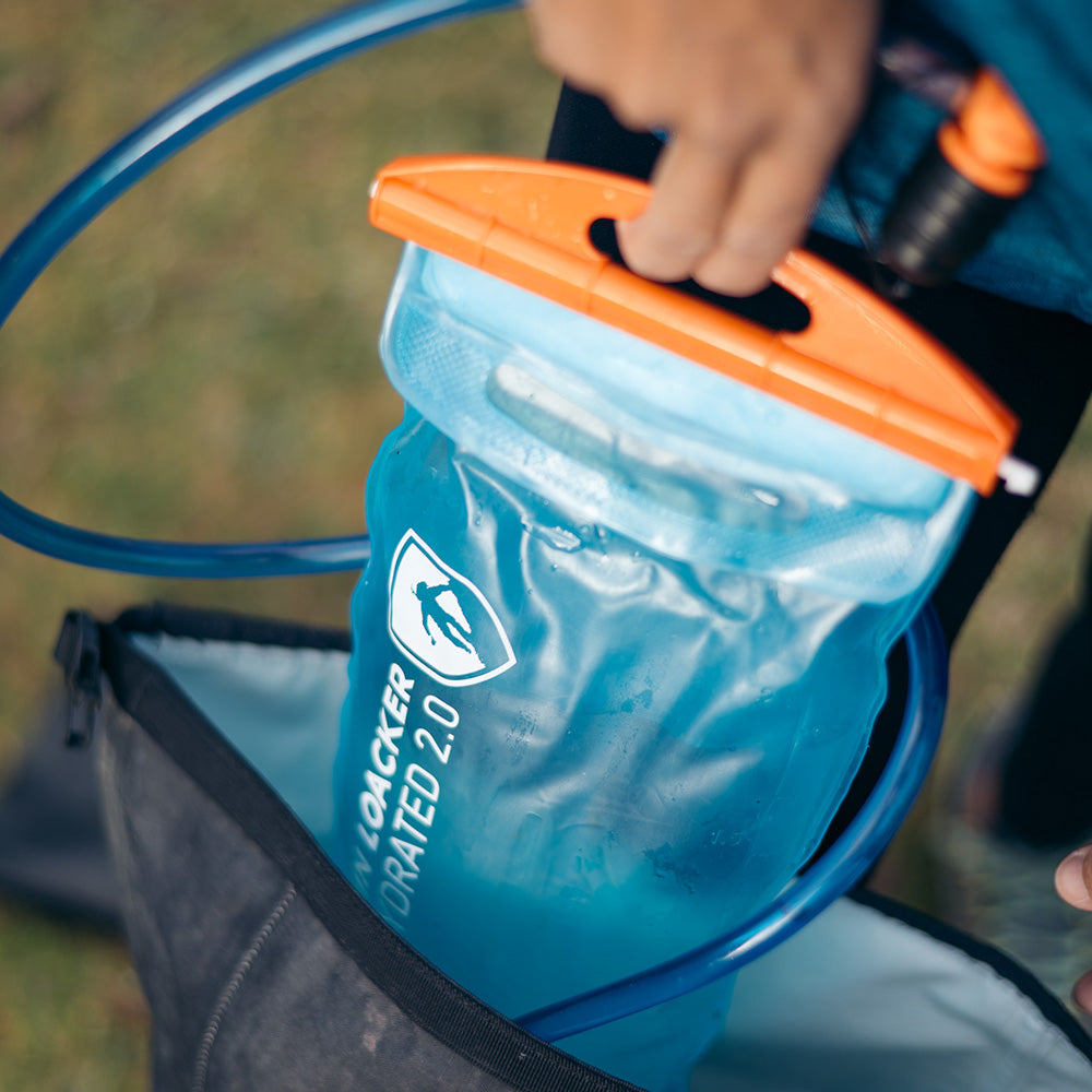 Trinkblase für Sommer + Winter: Nahaufnahme der blauen Wasserblase mit orangefarbener Verschlussklemme und Trinkschlauch, aus einem Rucksack gezogen, ideal für Outdoor-Sport.