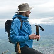 Person mit Alpin Loacker Sonnenhut mit UV Schutz Unisex, blauem Regenjacke und schwarzem Rucksack, steht auf Berg, hält Trekkingstöcke, überblickt Tal.