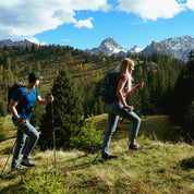 Zwei Wanderer, einer im Merino T-Shirt Damen 150g/m2, nutzen Trekkingstöcke auf einem grasbewachsenen Hang mit schneebedeckten Bergen im Hintergrund.
