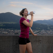 Frau mit Merino Coolmax T-Shirt Damen 150 g/m2 trinkt aus Trinkflasche, ideal für Outdoor-Aktivitäten, bietet Temperaturausgleich und Komfort.