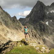 Hiker trägt Merino Coolmax T-Shirt Damen 150 g/m2 auf einem grasbewachsenen Felsvorsprung vor schneebedeckten Bergspitzen, ideal für Outdoor-Aktivitäten und Temperaturanpassung.