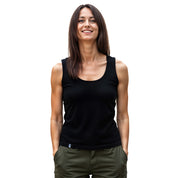 Junge Frau trägt Merino Tank Top Damen 150g/m2, ideal für Outdoor-Aktivitäten. Sie lächelt, trägt schwarze Tanktop und olivgrüne Hose, Hände in den Taschen.