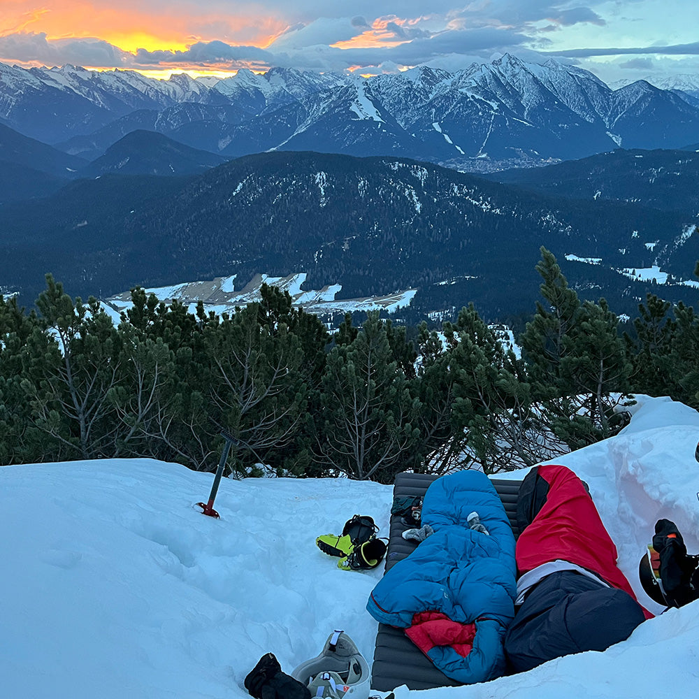 Biwaksack mit Membrane - NEU: Zwei Bergsteiger in einem winterlichen Biwak mit Alpin Loacker Biwaksäcken, umgeben von alpinem Kletterzubehör auf schneebedecktem Berghang bei Sonnenuntergang.