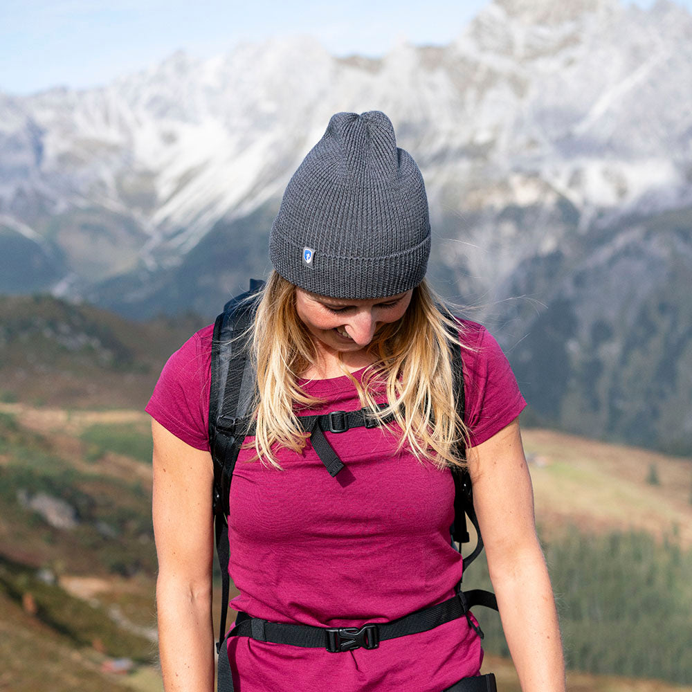 Person mit grauer Merino Mütze Herren & Damen Strick 230g/m², steht in Berglandschaft, trägt magenta T-Shirt und Wander-Rucksack. Ideal für Outdoor-Aktivitäten.
