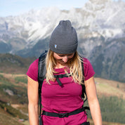 Person mit grauer Merino Mütze Herren & Damen Strick 230g/m², steht in Berglandschaft, trägt magenta T-Shirt und Wander-Rucksack. Ideal für Outdoor-Aktivitäten.