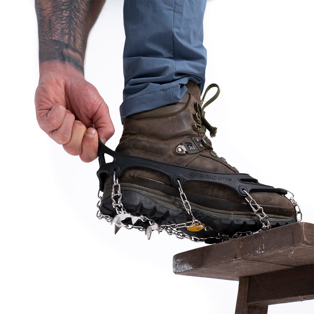 Hand zieht schwarzen Gummirahmen der Alpin Loacker Chain Pro 18 Grödel über robusten Wanderstiefel, um Metallketten und Spikes für sicheren Halt auf eisigem Terrain zu befestigen.