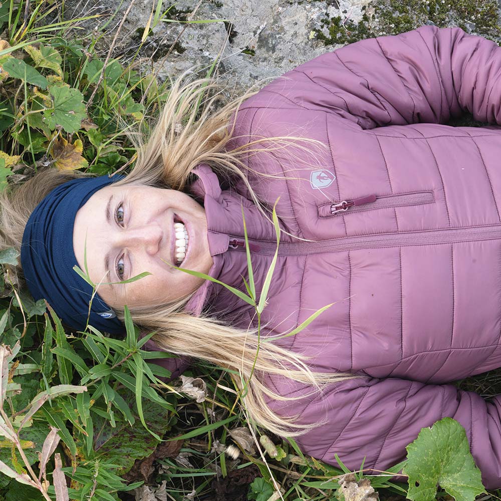 Person in mauver Jacke und Stirnband liegt lächelnd im Freien, trägt Merino Halstuch Herren & Damen, ideal für Outdoor-Aktivitäten bei kühlem Wetter, aus atmungsaktiver Merinowolle.