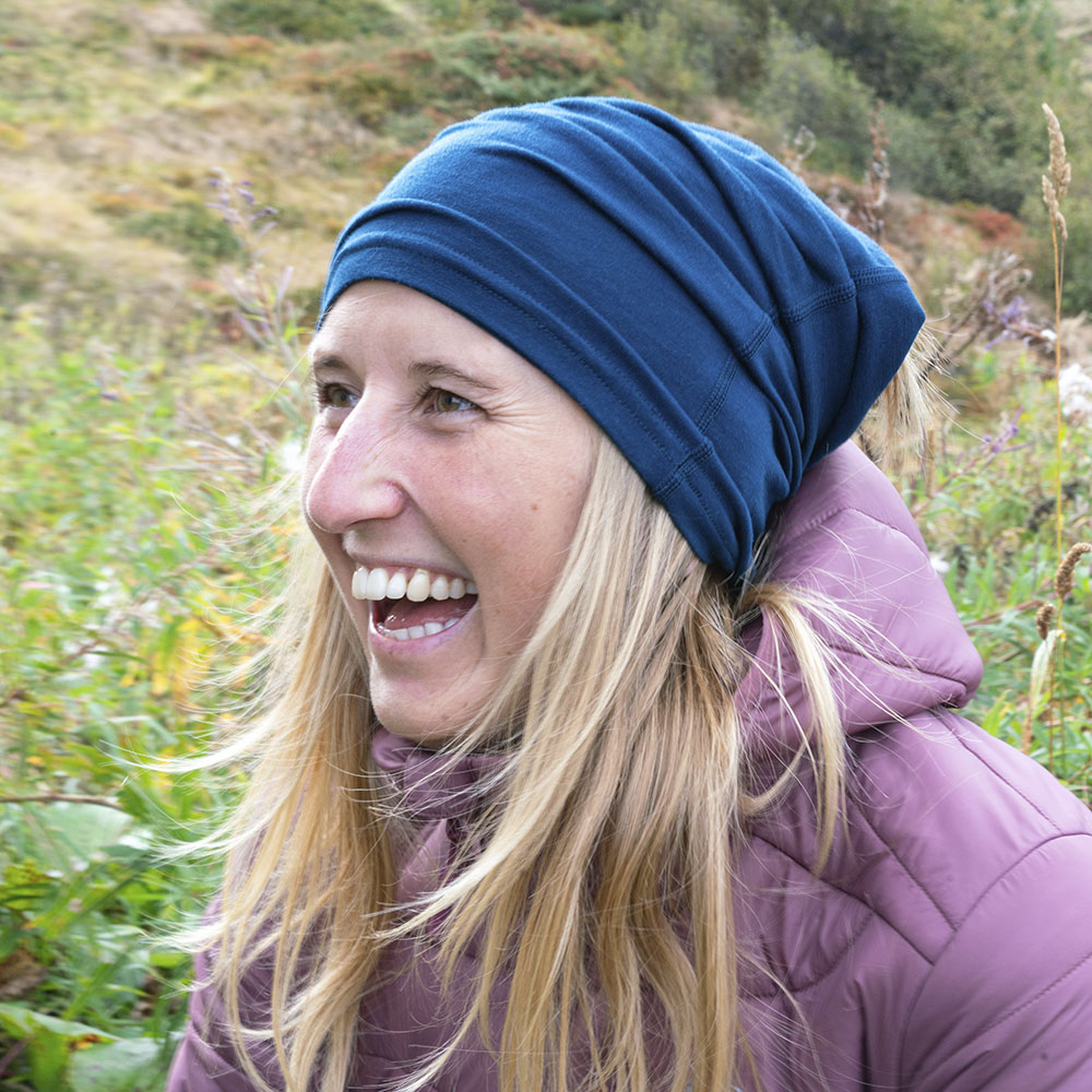 Person im Freien trägt blaues Beanie, lila Pufferjacke, lacht. Fokus auf Merino Halstuch Herren & Damen, ideal für Outdoor-Aktivitäten bei kühlem Wetter, atmungsaktiv, elastisch.
