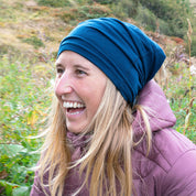 Person im Freien trägt blaues Beanie, lila Pufferjacke, lacht. Fokus auf Merino Halstuch Herren & Damen, ideal für Outdoor-Aktivitäten bei kühlem Wetter, atmungsaktiv, elastisch.