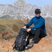 Mann mit blauem Fleece auf Felsen, neben ihm der Alpin Loacker 35-Liter Wanderrucksack, ideal für Outdoor-Abenteuer, mit Trekking-Ausrüstung und Edelstahlflasche.