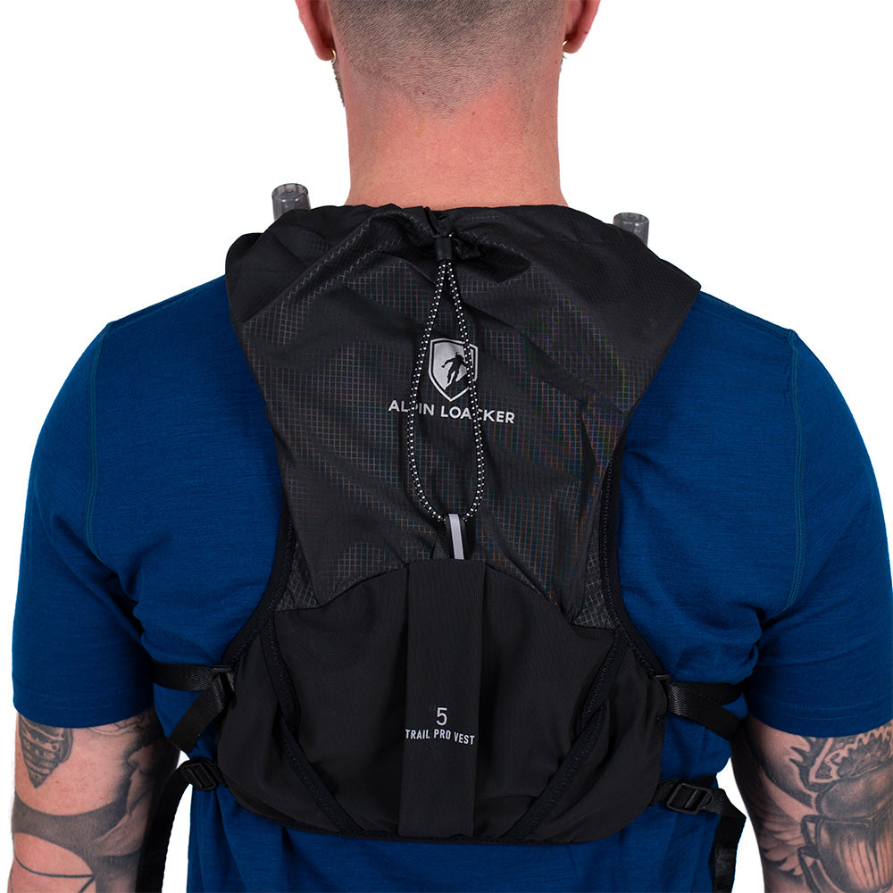 Laufweste Trail Pro 5 mit 2 Flaschen, schwarze Alpin Loacker Hydrationsweste mit Taschen, Kordelkapuze, ideal für Trailrunning, über blauem T-Shirt getragen.