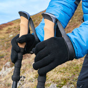 Person trägt schwarze Merino Handschuhe Damen & Herren, Classic, beim Halten von Trekkingstöcken. Perfekt für Outdoor-Aktivitäten, bieten sie Wärme und Geschicklichkeit.