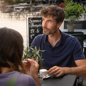Mann im Outdoor-Café trägt das Merino Polo Shirt Herren, genießt Kaffee. Ideal für Outdoor-Aktivitäten, atmungsaktiv, temperaturregulierend und geruchsresistent.