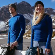 Merino Langarm Shirt Damen 230 g/m2: Paar in blauen Base-Layers vor verschneiten Bergen, bereit für Outdoor-Abenteuer mit Rucksäcken und Wintersportausrüstung.