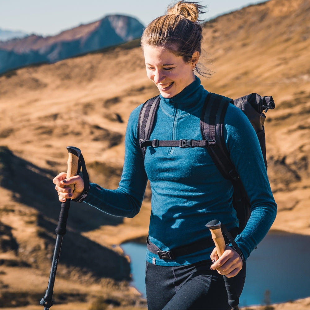 Frau mit blauem Top wandert mit Alpin Loacker Teleskop Wanderstöcke aus Carbon auf einem Bergpfad, umgeben von Hügeln und einem See im Hintergrund.