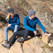 Zwei Wanderer pausieren auf einem felsigen Hang, der Mann trägt die Merino Mütze Herren & Damen Strick 230g/m² für optimalen Komfort und Temperaturregulierung.