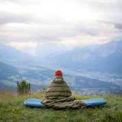 Person sitzt in UL Down Quilt 450g auf blauem Schlafpad, schaut über Tal. Ideal für Outdoor-Abenteuer mit leichtem, warmem Komfort bei kühlen Temperaturen.