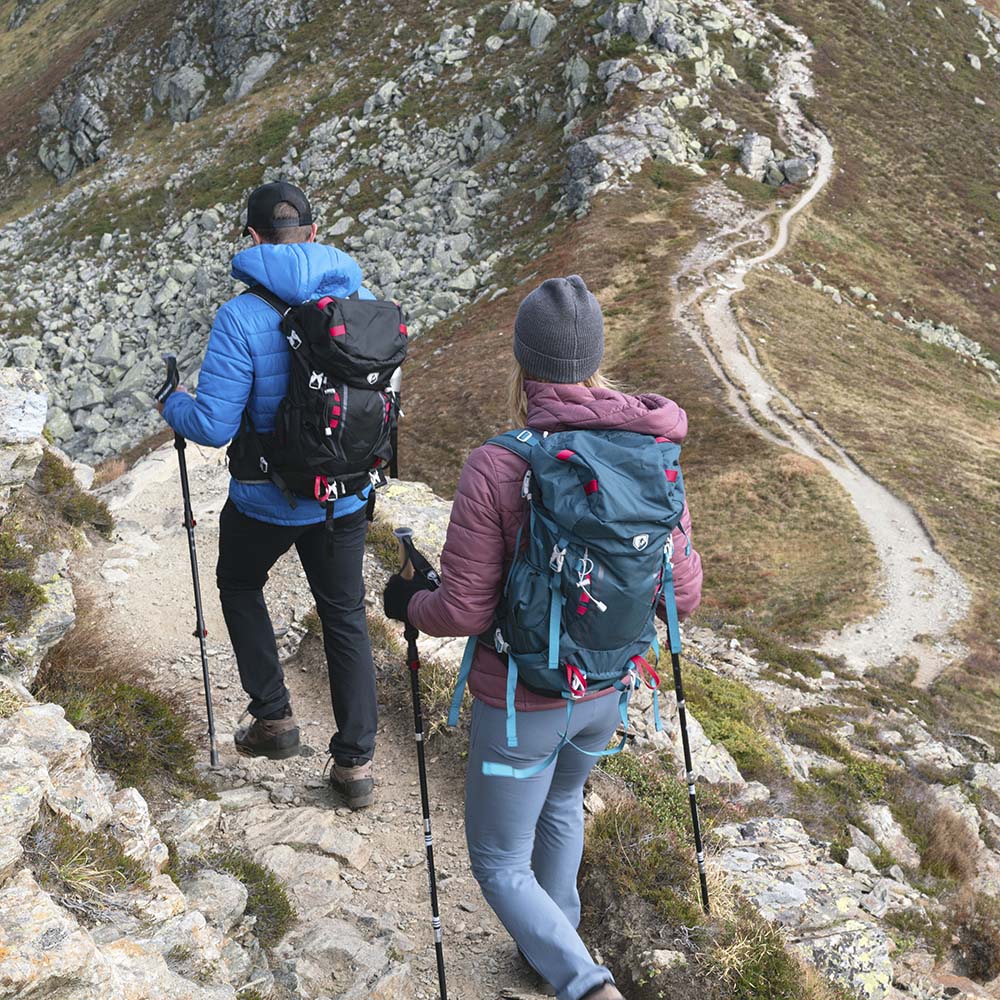 Zwei Wanderer mit dem 35-Liter-Wanderrucksack von Alpin Loacker auf einem steinigen Bergpfad, ausgestattet mit Trekkingstöcken und wetterfester Outdoor-Ausrüstung.