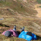 Zwei Personen auf einem Bergabhang tragen blaue und pinke Jacken, genießen die Aussicht, tragen leichte Merino Wandersocken Herren & Damen von Alpin Loacker für optimalen Wanderkomfort.