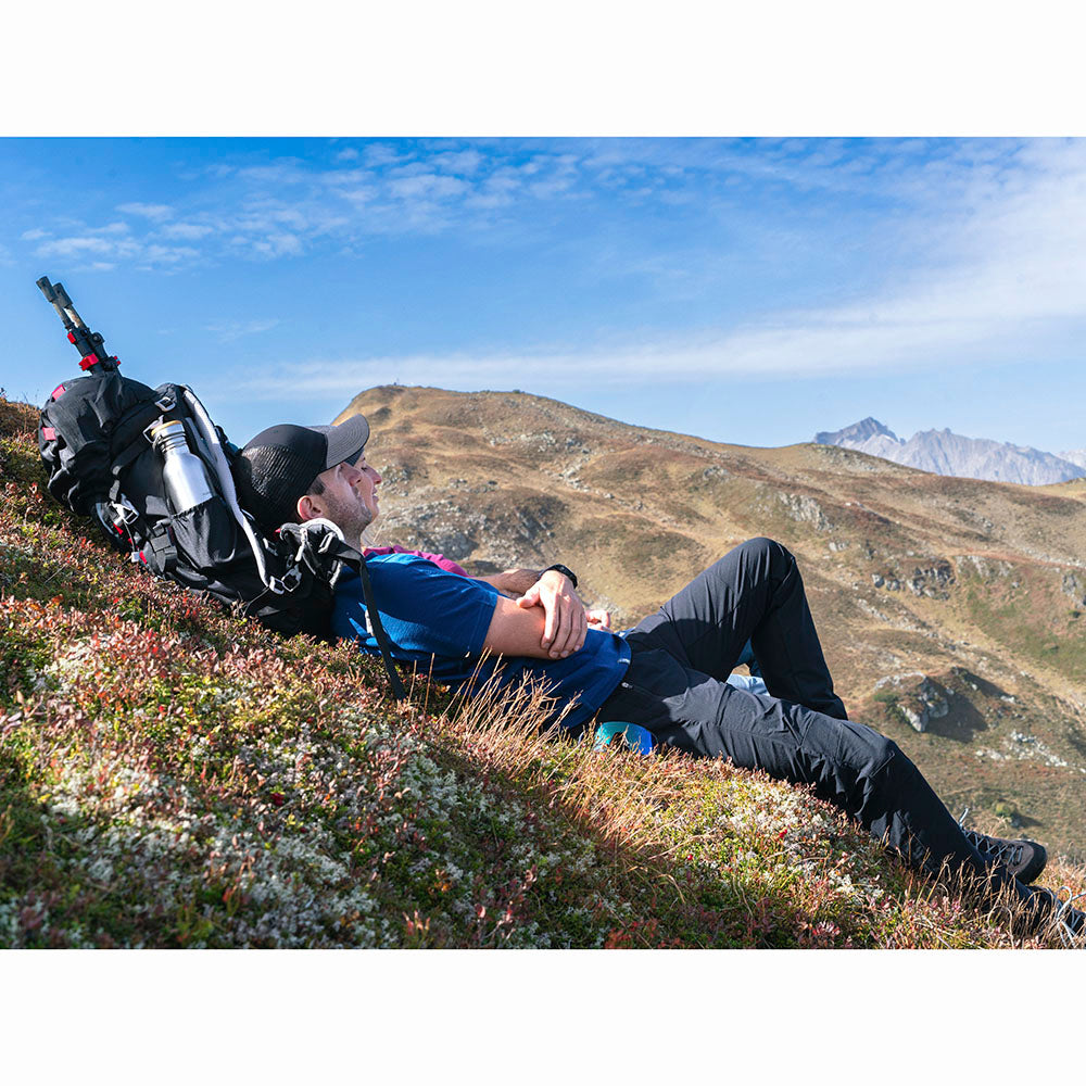 Mann entspannt auf Bergwiese, nutzt großen Rucksack als Kissen. Trägt Softshell Wanderhose Herren, ideal für Trekking mit Komfort und Bewegungsfreiheit.