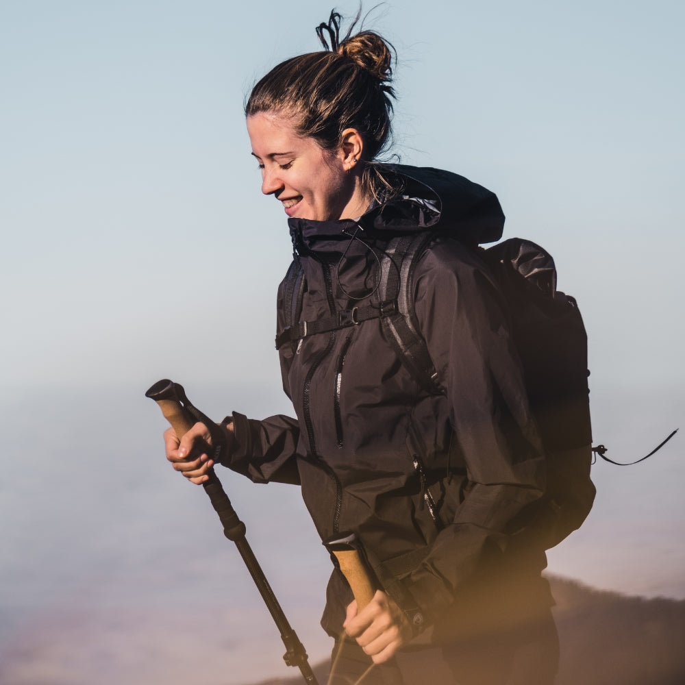 Person beim Wandern mit 3 Lagen Damen Hardshell Regenjacke, Trekkingstöcken und Rucksack, genießt die Aussicht. Jacke bietet Schutz bei jedem Wetter.