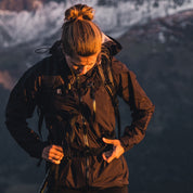 Person beim Wandern mit der 3 Lagen Damen Hardshell Regenjacke, justiert den Hüftgurt des Rucksacks. Wetterfeste, atmungsaktive Jacke für Outdoor-Aktivitäten. Ideal für wechselnde Bedingungen.