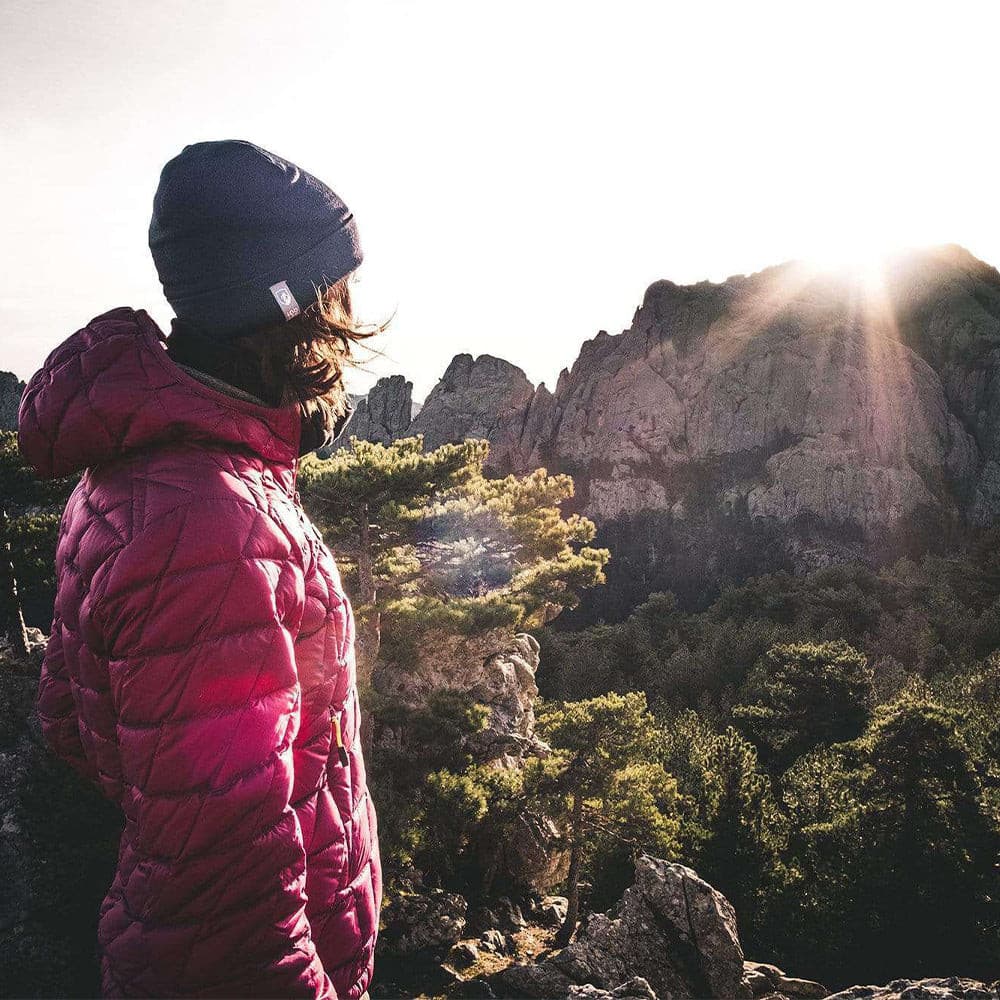 Person mit Merino Mütze Herren & Damen 230g/m2 blickt von Felsen über bewaldetes Gebirge im Sonnenaufgang. Ideal für Outdoor-Abenteuer, bietet Schutz und Komfort.