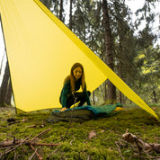 Person richtet unter einem gelben Tarp 500 im Wald einen Schlafplatz ein. Das leichte Wingtarp schützt bei Outdoor-Abenteuern vor Wetter.