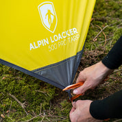 Nahaufnahme des Tarp 500 - das leichte Wingtarp von ALPIN LOACKER, aufgespannt im Wald, mit verstärkter Ecke und Person, die einen orangenen Hering befestigt.