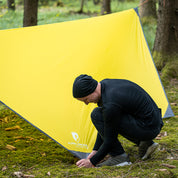 Person befestigt das Tarp 500 auf waldboden, ein leichtes, vielseitiges Wingtarp für Outdoor-Abenteuer, ideal als Regen- oder Sonnenschutz für 1-2 Personen.
