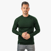Mann trägt Merino Langarm Shirt Herren 150 g/m2 in Dunkelgrün, passend für Outdoor-Aktivitäten, kombiniert mit grauen Jeans vor einem neutralen Hintergrund.