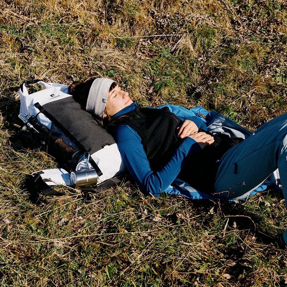 Frau entspannt auf einer Wanderung, trägt die Merino Weste Damen 270g/m2 als Midlayer, nutzt Rucksack als Kissen, Metallbecher am Rucksack befestigt.
