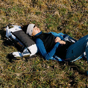 Frau entspannt auf einer Wanderung, trägt die Merino Weste Damen 270g/m2 als Midlayer, nutzt Rucksack als Kissen, Metallbecher am Rucksack befestigt.