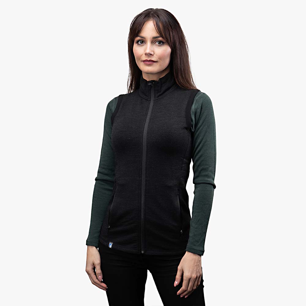 Frau trägt Merino Weste Damen 270g/m² über Langarmshirt, ideal als Midlayer für Outdoor-Aktivitäten. Sportlicher Schnitt, mit Seitentaschen und hohem Kragen.