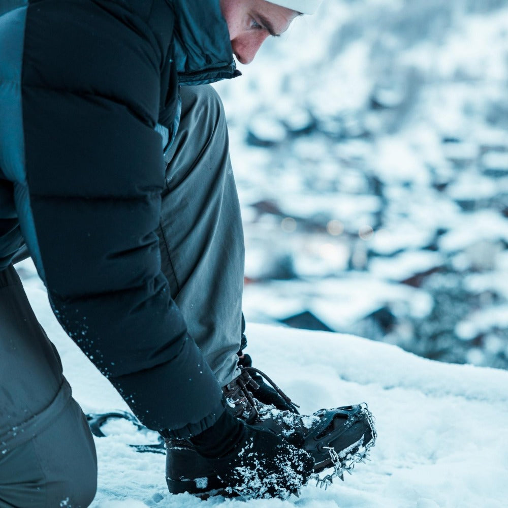 Person legt Chain Pro 18 Grödel mit 18 Edelstahl-Spikes über Winterstiefel an, bereit für eisiges Gelände. Perfekt für Winterwanderungen, Trekking und Bergtouren.