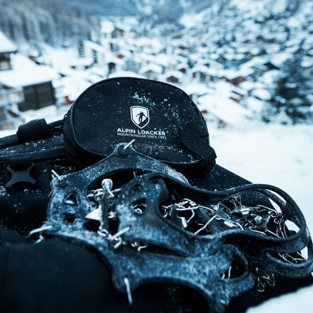 Alpin Loacker Chain Pro 18 Grödel auf schwarzem Tragebeutel, mit Schnee bedeckt, ideal für sicheren Halt auf winterlichen Wegen.