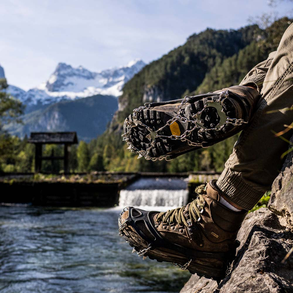 Hiker-Boots mit Alpin Loacker Chain Pro 18 Grödeln auf Felsen, umgeben von Berglandschaft. Edelstahl-Spikes bieten sicheren Halt auf Schnee und Eis.
