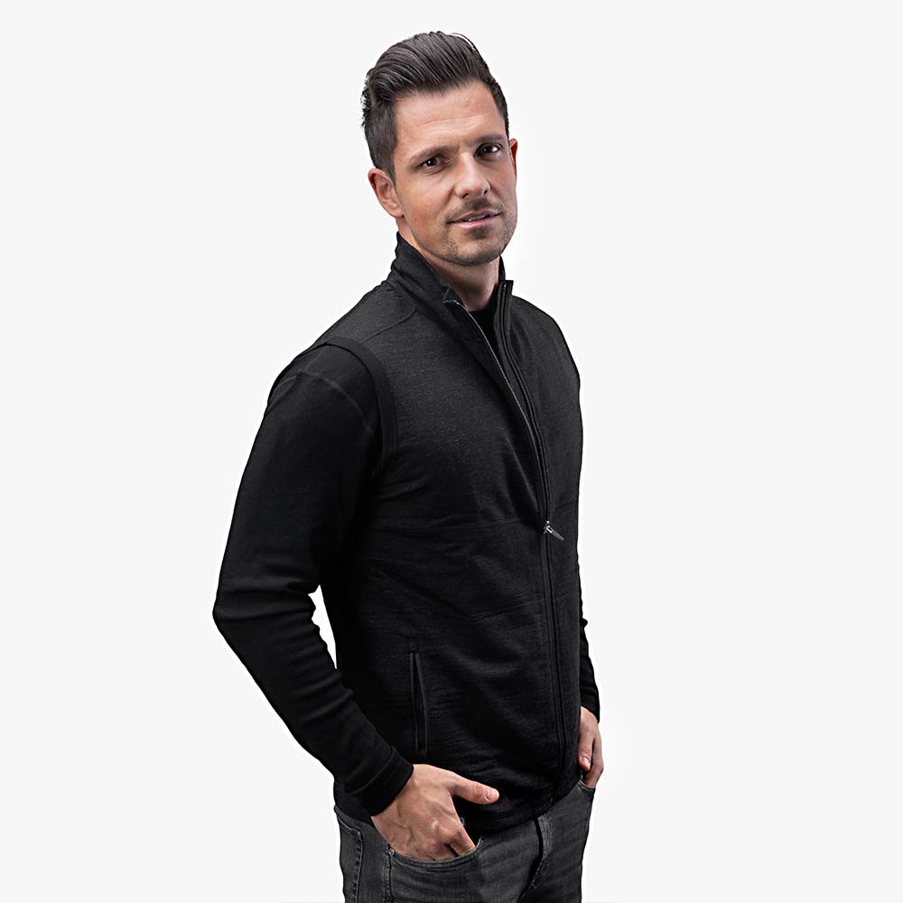 Mann trägt Merino Weste Herren 270 g/m2 über schwarzem Shirt, kombiniert mit dunklen Jeans. Perfekt als Midlayer für Outdoor-Aktivitäten und Freizeit.
