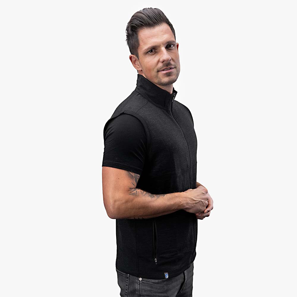 Mann trägt die Merino Weste Herren 270 g/m², eine ärmellose, schwarze Weste mit Zipper und Hüfttaschen, ideal für Outdoor-Aktivitäten und Alltag.