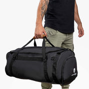 Person mit olivgrüner Cargo-Hose trägt eine schwarze Reisetasche, Sporttasche & Duffel Bag von Alpin Loacker mit Logo und seitlichen Reißverschlusstaschen.